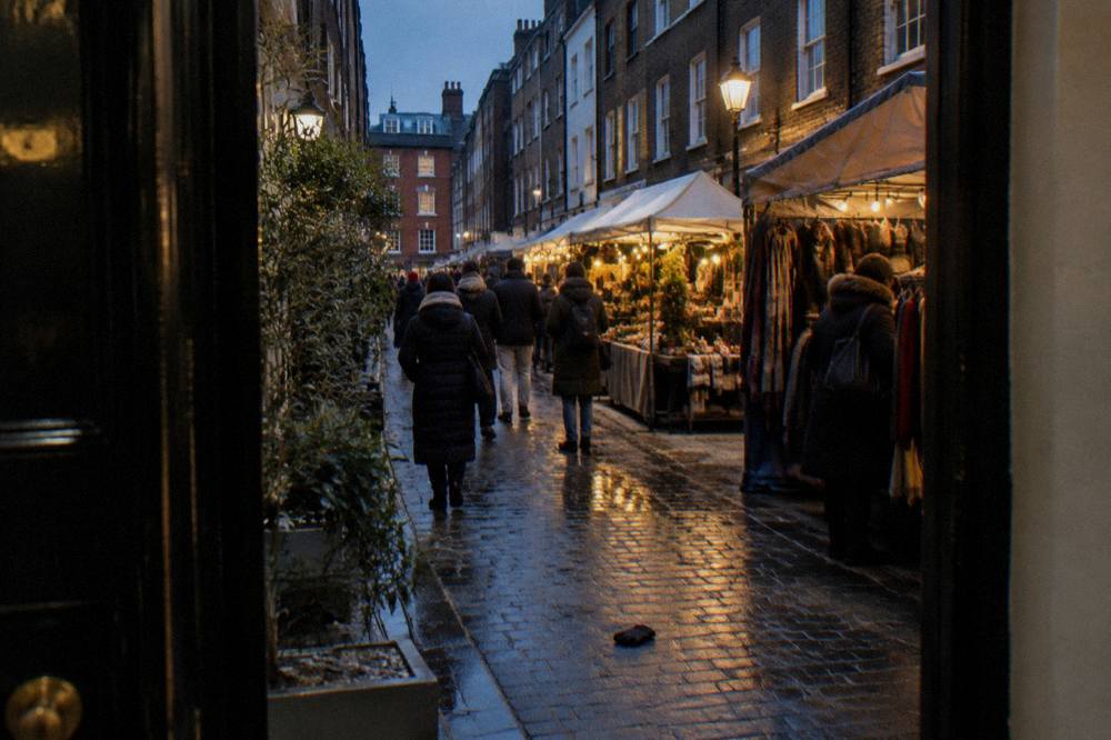 Winter shoppers in coats and scarves walking through a Christmas market with lit stalls in the early evening
