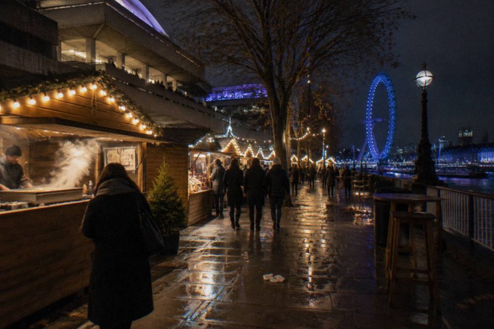 Crowded festive market stalls under string lights beside the Thames at dusk