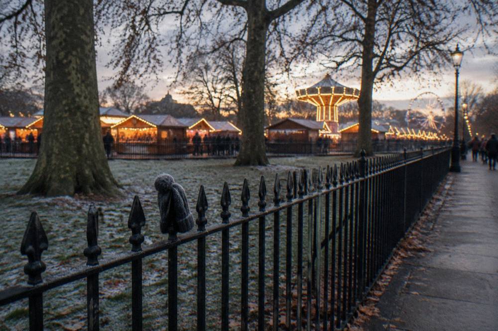 Row of lit Christmas market chalets beside a large park at dusk
