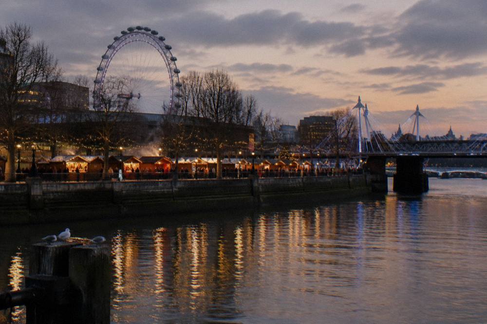 Illuminated Christmas market stalls along the South Bank at dusk with string lights and a lit bridge beyond