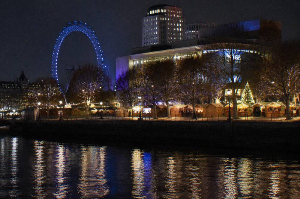 Festive stalls and illuminated trees along a riverside promenade with city lights reflected in the water
