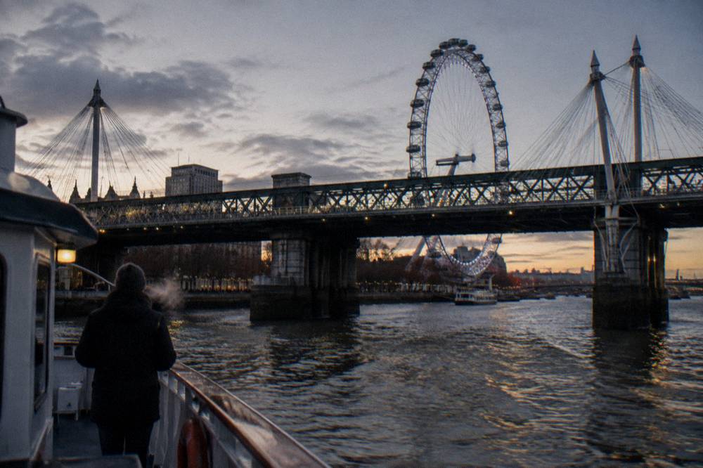 A cruise boat passing under an illuminated bridge on the Thames in the early evening