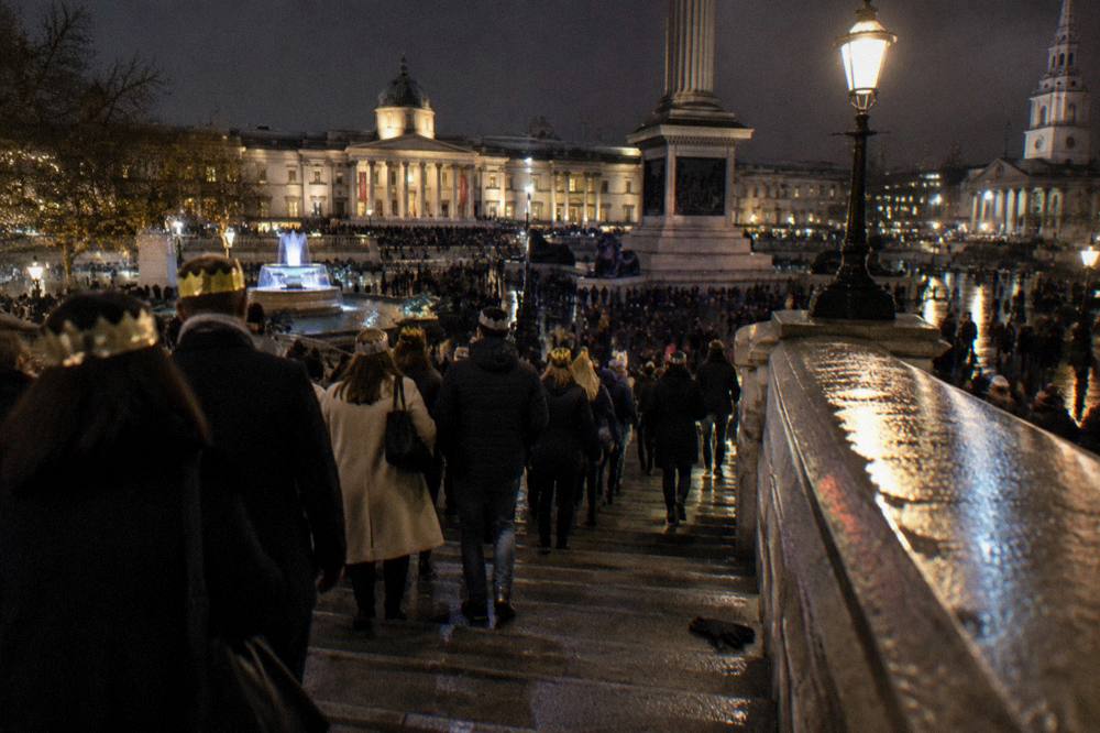 A crowded London bridge at night with crowds walking and city lights behind