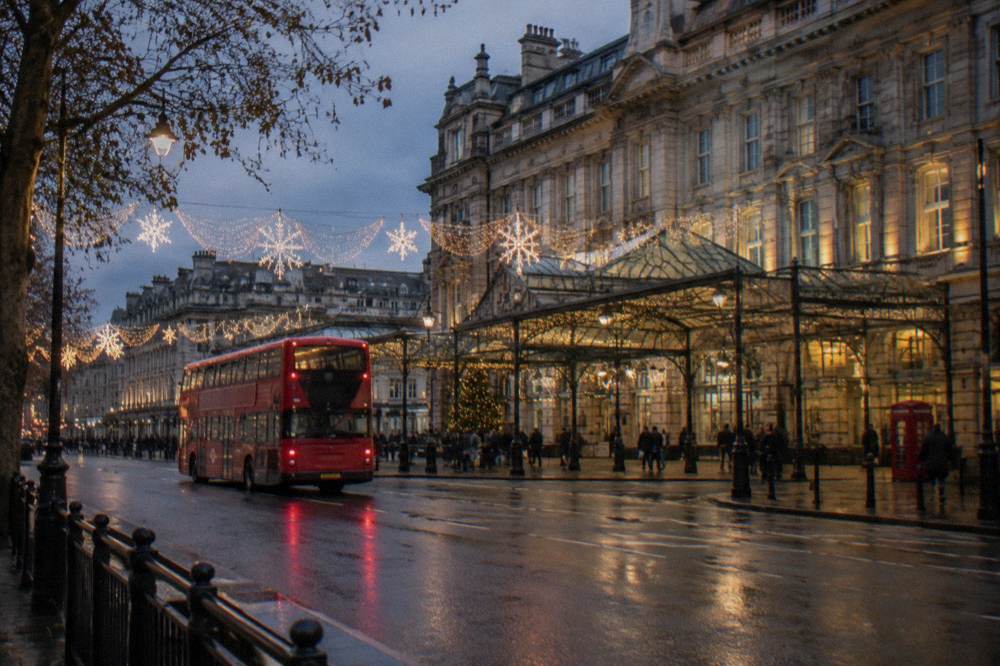 A red double-decker bus passing under glowing Christmas lights on a London street at dusk