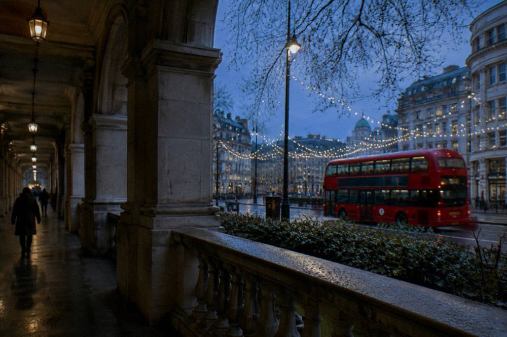 A red double-decker bus passing under glowing Christmas lights on a London street at dusk