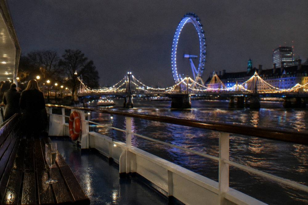 A cruise boat on the River Thames at night with lit bridges and a ferris wheel in the background