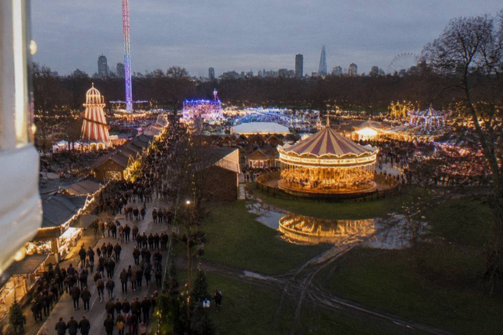 Crowds walking between illuminated fairground rides and food stalls in a large park at dusk