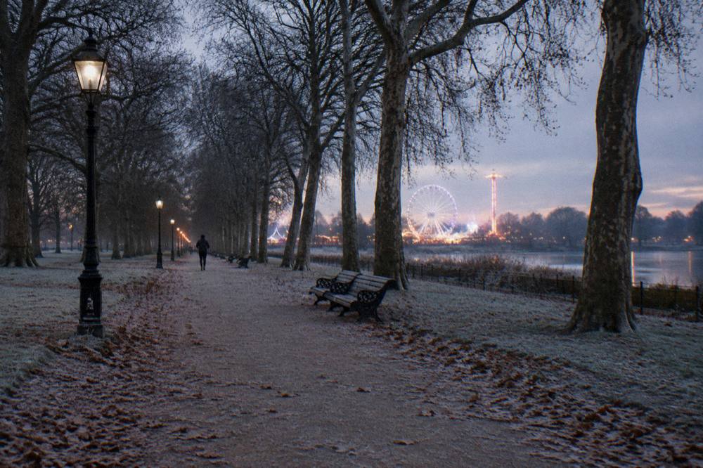 Frosty tree-lined path in a London park with distant fairground lights glowing at twilight